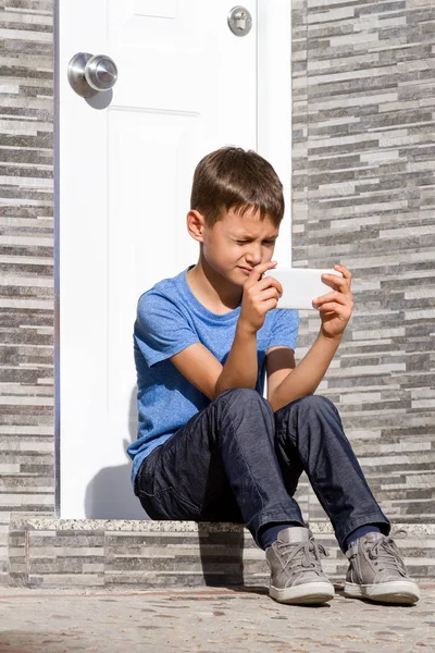 Menino com telefone celular sentado na escada em frente a uma porta — Fotografia de Stock