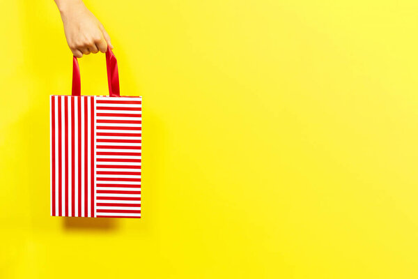 Рука держит красный полосатый подарок мешок на желтом фоне. Концепция шопинга, отдыха, пожертвований
