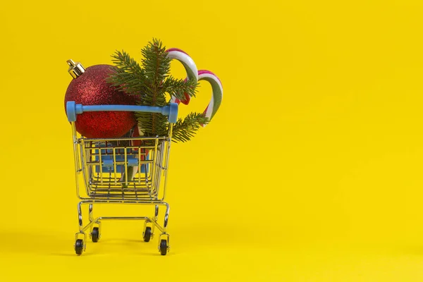 Mini carrinho de compras com decoração de Natal bugigangas, ramo de abeto e bengalas de doces no fundo amarelo — Fotografia de Stock