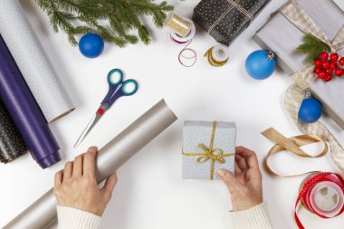 Noel hediyesi paketi. Kadın ellerine tepeden bakıldığında hediye kutuları beyaz masanın üzerinde köknar ağacı dalları, dekorasyonlar ve kurdelelerle sunuluyor.