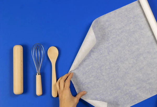 Woman hands roll baking paper. Wooden spoon, rolling pin and balloon whisk on blue background. Top view — 스톡 사진