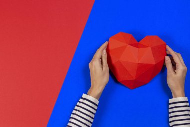 Mavi ve kırmızı arka planda kırmızı çokgen kalp tutan kadın eller. Üst görünüm