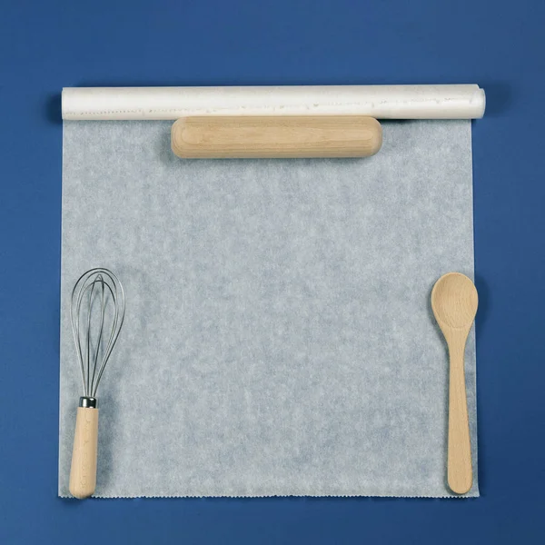 Wooden spoon, rolling pin and balloon whisk over white baking paper on blue background. Top view, flat lay — Stock Photo, Image