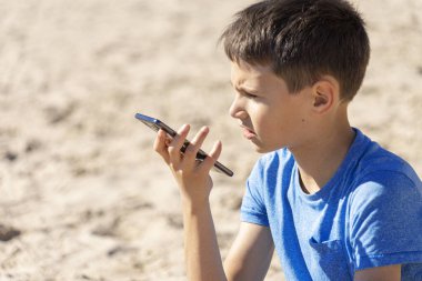 Genç bir çocuk, akıllı bir telefon ses tanıma işlevini online olarak kullanıyor, cep telefonunu kullanarak sahilde oturan bir sesli mesaj gönderiyor.
