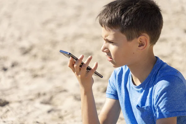 Menino adolescente usando uma função de reconhecimento de voz de telefone inteligente on-line, usando telefone celular para enviar uma mensagem de voz sentada na praia — Fotografia de Stock