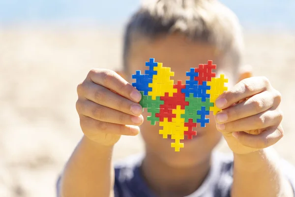 Chłopiec trzyma kolorowe serce układanki przed twarzą. Światowa koncepcja Dnia Świadomości Autyzmu — Zdjęcie stockowe