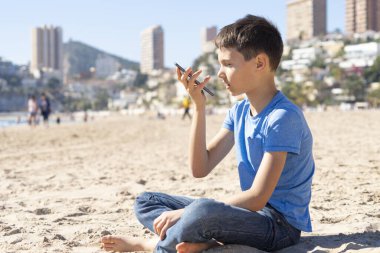 Genç bir çocuk akıllı bir telefon ses tanıma işlevini online olarak kullanıyor, cep telefonunu kullanarak şehir plajında oturan bir sesli mesaj gönderiyor.