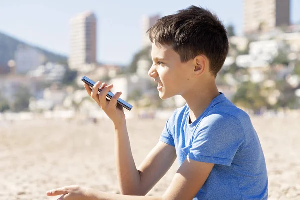 Menino adolescente usando uma função de reconhecimento de voz de telefone inteligente on-line, usando telefone celular para enviar uma mensagem de voz sentada na praia da cidade — Fotografia de Stock