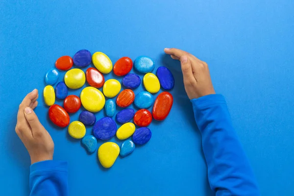 World autism awareness day concept. Child hands with heart made of blue yellow red color stones over light blue background — Stock Photo, Image