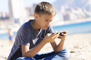 Şehir plajında telefonuyla oturan bir genç. Çocuk sosyal medyaya odaklanmış, oyunlar oynuyor, sohbet ediyor, okuyor, video izliyor.