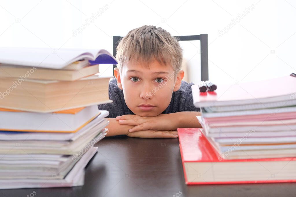 Tired disappointed schoolboy sitting among pile of books, textbooks, school exercise books on his desk at home