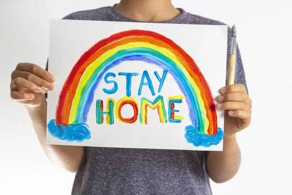 Niño sosteniendo dibujo con arco iris con palabras Quédese en casa. Campaña de medios sociales para la prevención del coronavirus — Foto de Stock