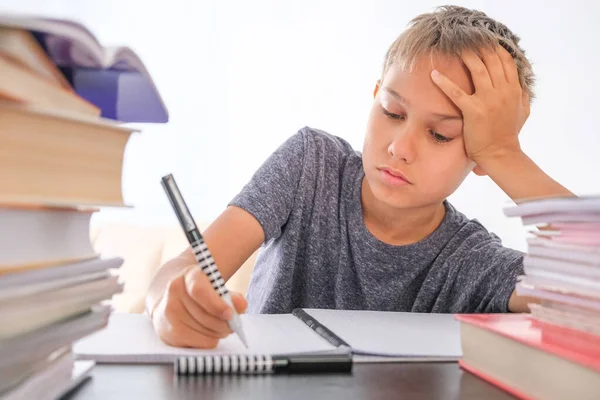 机の上の本、教科書、学校の運動本の山の間に座って、宿題をしている学童。教育、オンライン距離学習、自宅での隔離中のホームスクール — ストック写真