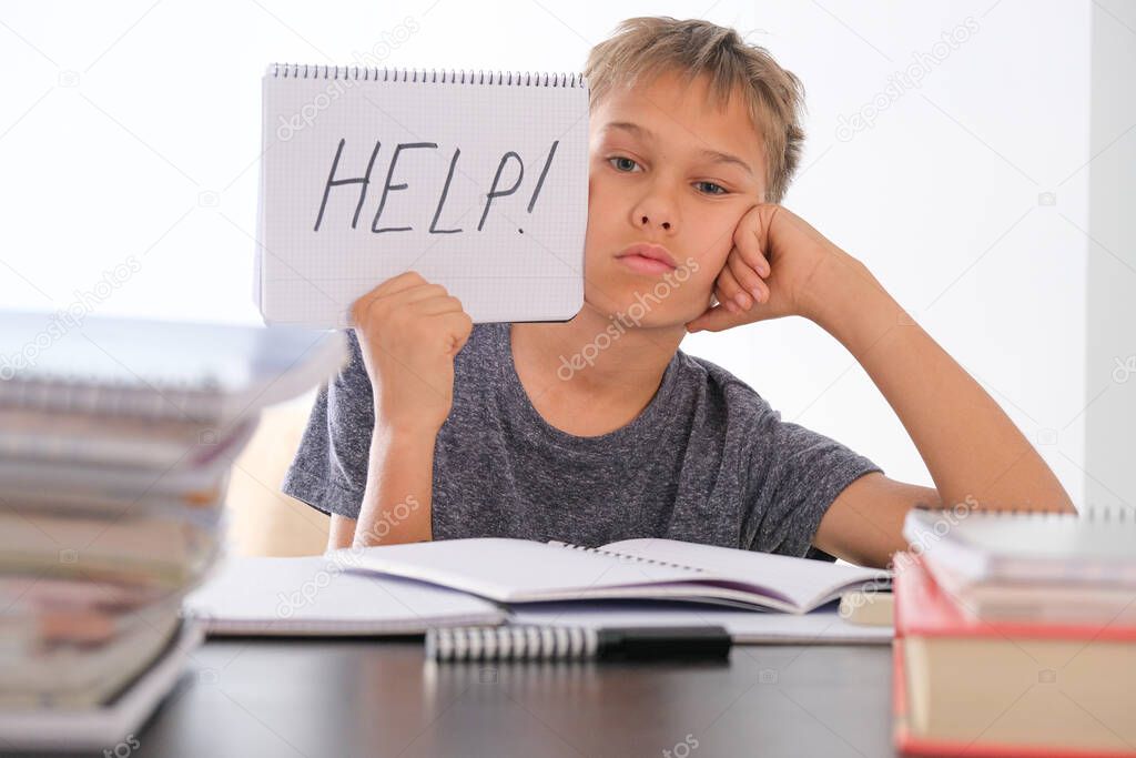 Upset tired preteen child sitting at the table, doing his homework among pile of books. Word Help is written on open notebook. Learning difficulties, school, education concept