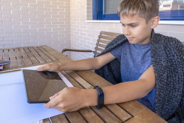 Çocuk dijital tablet bilgisayar ve not defterleriyle öğreniyor. Okul çocuğu balkonda ödev yapıyor. Çevrimiçi öğrenim, uzaktan eğitim, teknoloji, ev konseptinde kalın
