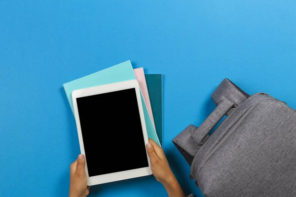 Kid hands with tablet computer, notebooks, books and gray backpack over light sky blue background — Stock Photo, Image