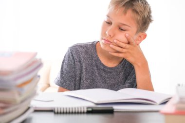 Öğrenci çocuk kitap yığınlarının arasında oturuyor, ders kitapları, çalışma kitapları masasında ve ödevlerini yapıyor. Eğitim, çevrimiçi öğrenme, evde uzaktan öğrenme