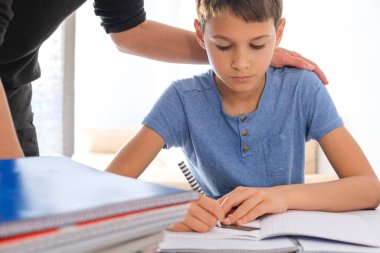 Çocuk elinde bir sürü defterle masada oturuyor ve ödev yapıyor. Annem ona yardım ediyor. Öğrenme zorluğu, evde öğrenme, eğitim, motivasyon kavramı
