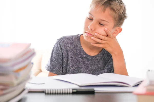 Iskolásfiú ül egy halom könyv, tankönyvek, iskolai füzetek között az asztalán, és házi feladatot csinál. Oktatás, online tanulás, távoktatás otthon — Stock Fotó