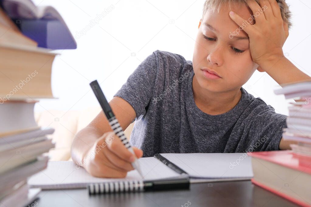 Schoolboy sitting among pile of books, textbooks, school exercise books on his desk and doing homework. Education, online distance learning, homeschooling during quarantine at home