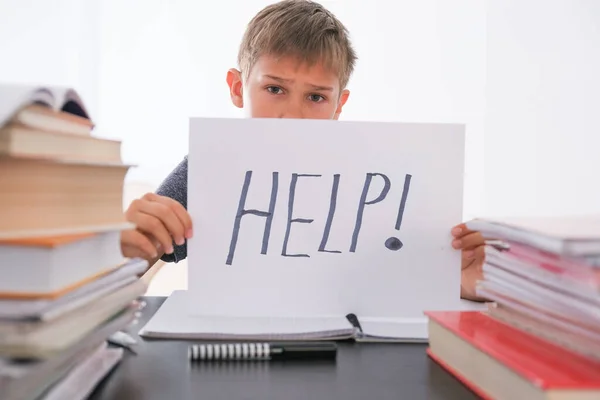 闷闷不乐的男孩坐在桌旁，手里拿着许多书，手里拿着写着"帮助"的纸。学习困难、教育观念. — 图库照片