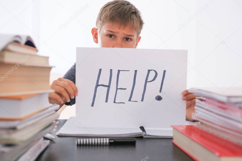 Sad tired frustrated boy sitting at the table with many books and holding paper with word Help. Learning difficulties, education concept.