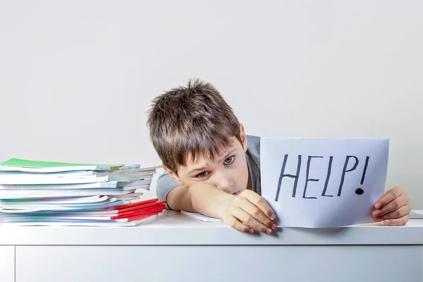 Triste criança frustrada cansado sentado à mesa com muitos livros e segurando papel com a palavra Ajuda. Dificuldades de aprendizagem, educação, aprendizagem online, escola em casa — Fotografia de Stock