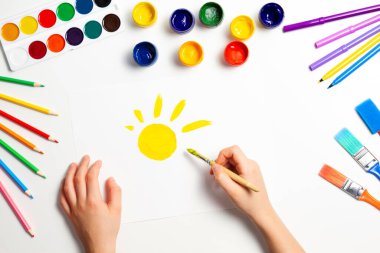 Çocuk elleri sarı güneşi boyuyor. Resim malzemeleriyle beyaz masaya üst görünüm