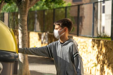 Koruyucu maskeli ve eldivenli çocuk boş plastik şişeyi dışarıdaki apartmanın yakınındaki geri dönüşüm kutusuna atıyor.