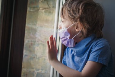 Küçük bir çocuk, tıbbi bir maskeyle pencereden dışarı bakıyor.