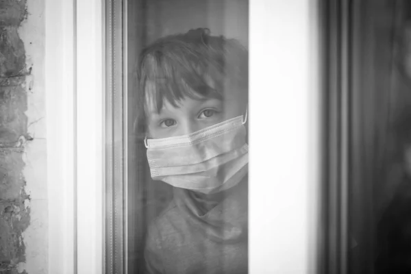 Un niño pequeño mira por la ventana con una máscara médica — Foto de Stock