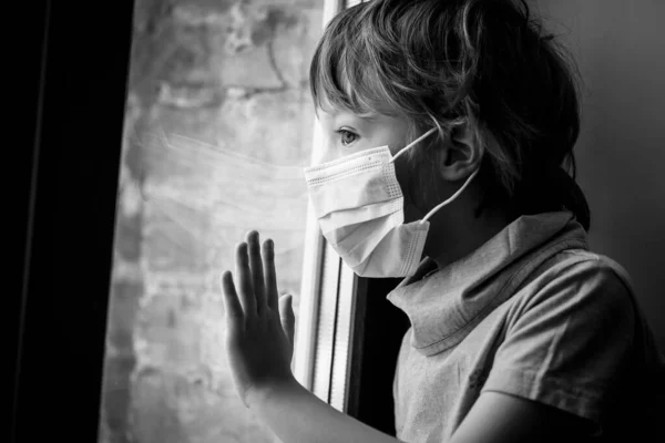 Un niño pequeño mira por la ventana con una máscara médica — Foto de Stock