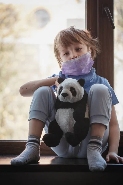 Niño triste chico se sienta en un alféizar de la ventana en una máscara médica — Foto de Stock