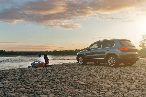 Portrait de couple heureux de jeunes adultes avec chien en voyage. Homme assis sur plaid avec une femme et regardant le coucher du soleil près de la rivière. Concept de pique-nique extérieur . — Photo