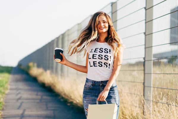 Piękna kobieta spaceru z kawy i torba na zakupy w biały T-shirt z słowo "mniej" — Zdjęcie stockowe