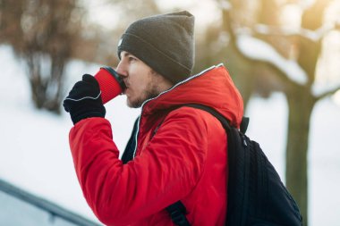 Genç adam gri şapka, kırmızı ceket ve siyah sırt çantası park ve sıcak kahve fincan holding parlak kış gün boyunca yürüyüş