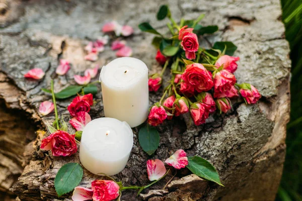 White candles with roses and green leaves on brown wooden background. Romantic atmosphere outdoors
