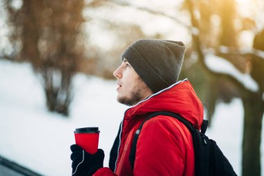 Genç adam gri şapka, kırmızı ceket ve siyah sırt çantası park ve sıcak kahve fincan holding parlak kış gün boyunca yürüyüş