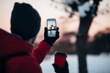 Gri ve kırmızı ceket, elinde fotoğraf smartphone ağaç üzerinde günbatımında kış parkta kahve fincan genç adam. Arkadan Görünüm