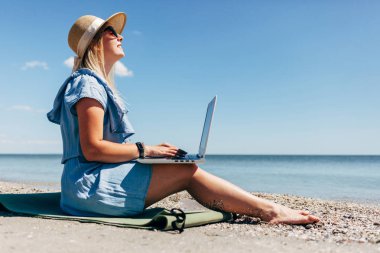 Arka planı denizin kucağında dizüstü bilgisayar ile sahilde oturan mavi elbise giyen genç kadın