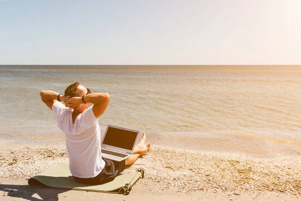 Genç adam bir beyaz T-shirt sahilde deniz arka plan üzerinde bir dizüstü bilgisayar ile oturur