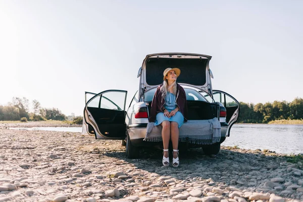 Young traveling woman in a blue dress is sitting by the car by the river — Stock Photo, Image