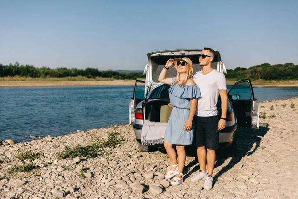 Feliz viajero pareja de pie cerca de coche abierto tronco y ver el amanecer —  Fotos de Stock