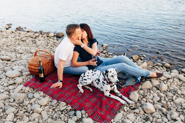 Portrait of happy young adult couple with dog on roadtrip. Man sitting on plaid with woman. Outdoor picnic concept.