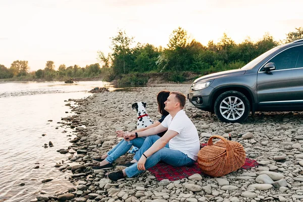 Portrait de couple heureux de jeunes adultes avec chien en voyage. Homme assis sur plaid avec une femme. Concept de pique-nique extérieur . — Photo