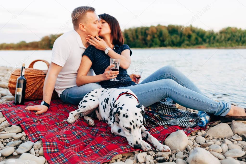 Portrait of happy young adult couple with dog on roadtrip. Man sitting on plaid with woman. Outdoor picnic concept.