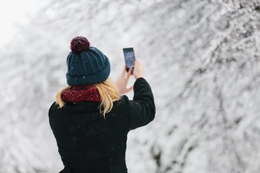 Siyah palto ve kırmızı örme eşarp genç Beyaz sarışın kız kış parkta smartphone ile yürür.