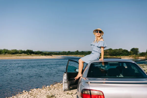 Молодая путешествующая женщина в синем платье сидит у машины у реки — стоковое фото