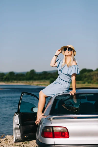 Молодая путешествующая женщина в синем платье сидит у машины у реки — стоковое фото