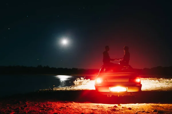 Happy traveler couple sitting on roof of the car and watching moonlight